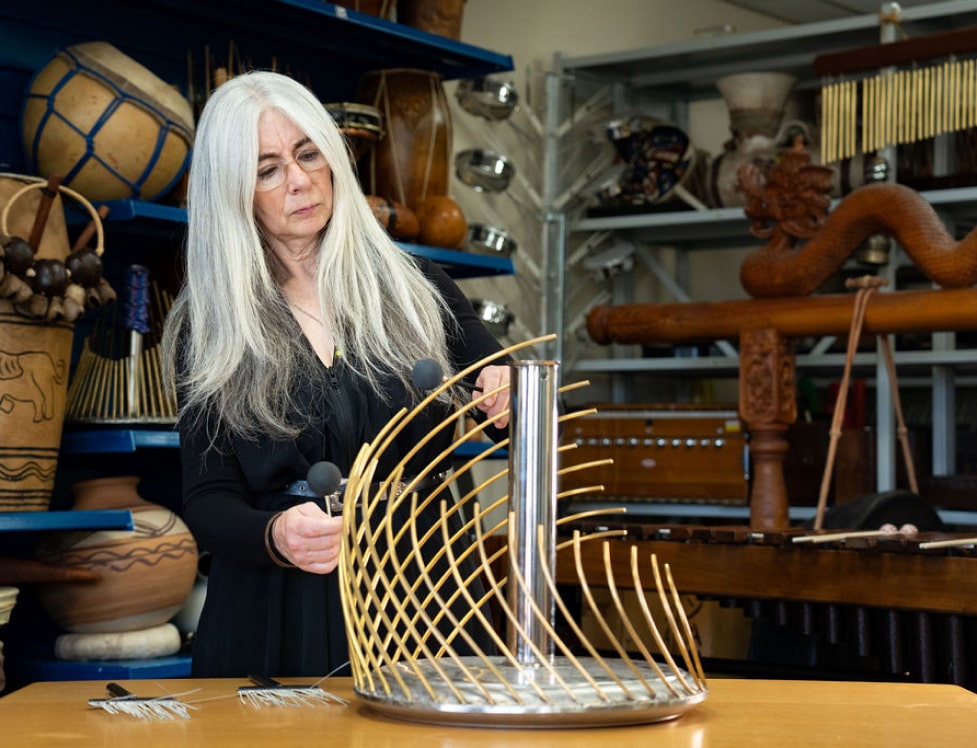 Evelyn stood, playing a Hyperstellar Sailophone, which is a unique type of Waterphone, as it rests on a table, both of which belong to The Evelyn Glennie Collection.