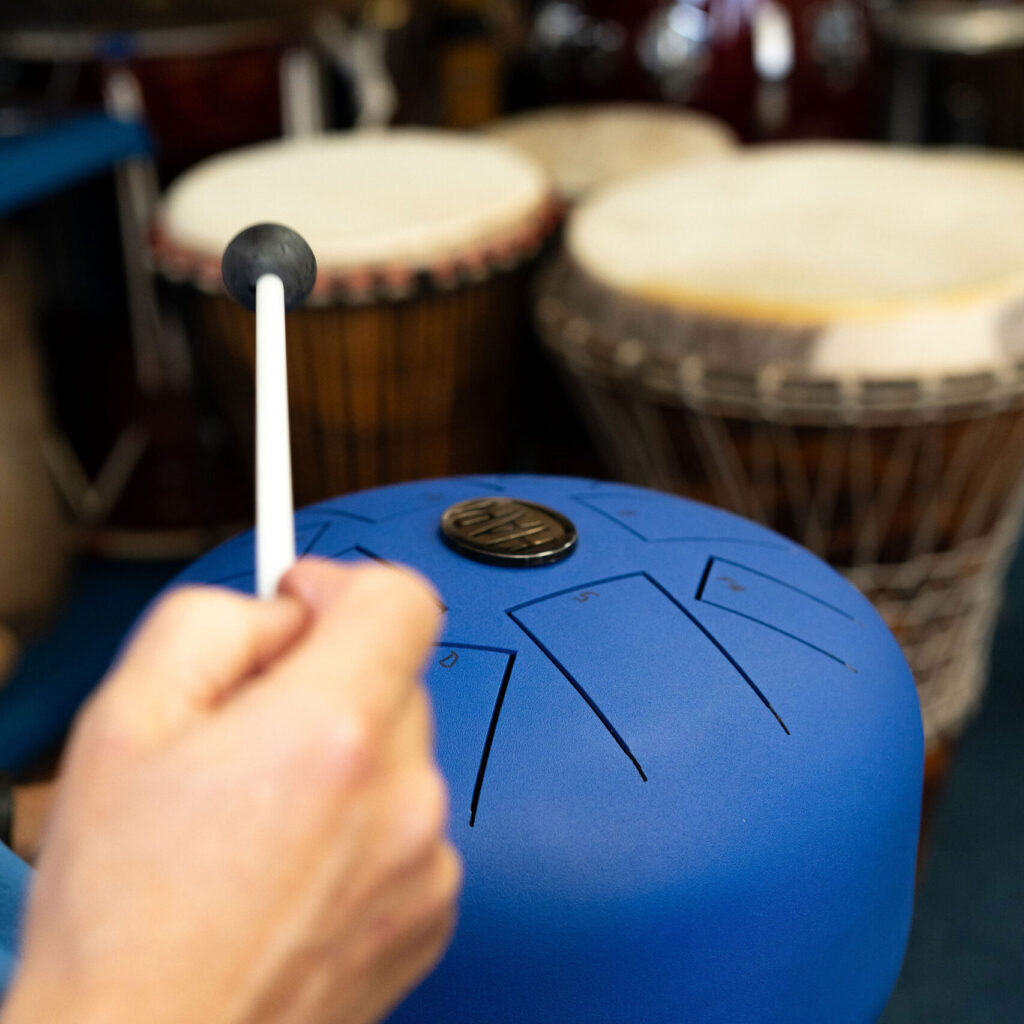 A participant striking a blue Hapi drum, with three Djembes out of focus in the background, during a 'Who's Listening' workshop.