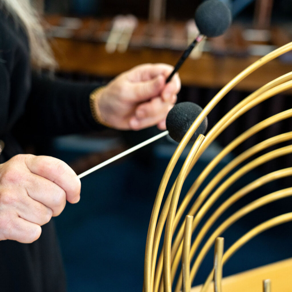 Evelyn stood, playing a Hyperstellar Sailophone, which is a unique type of Waterphone, as it rests on a table, both of which belong to The Evelyn Glennie Collection.